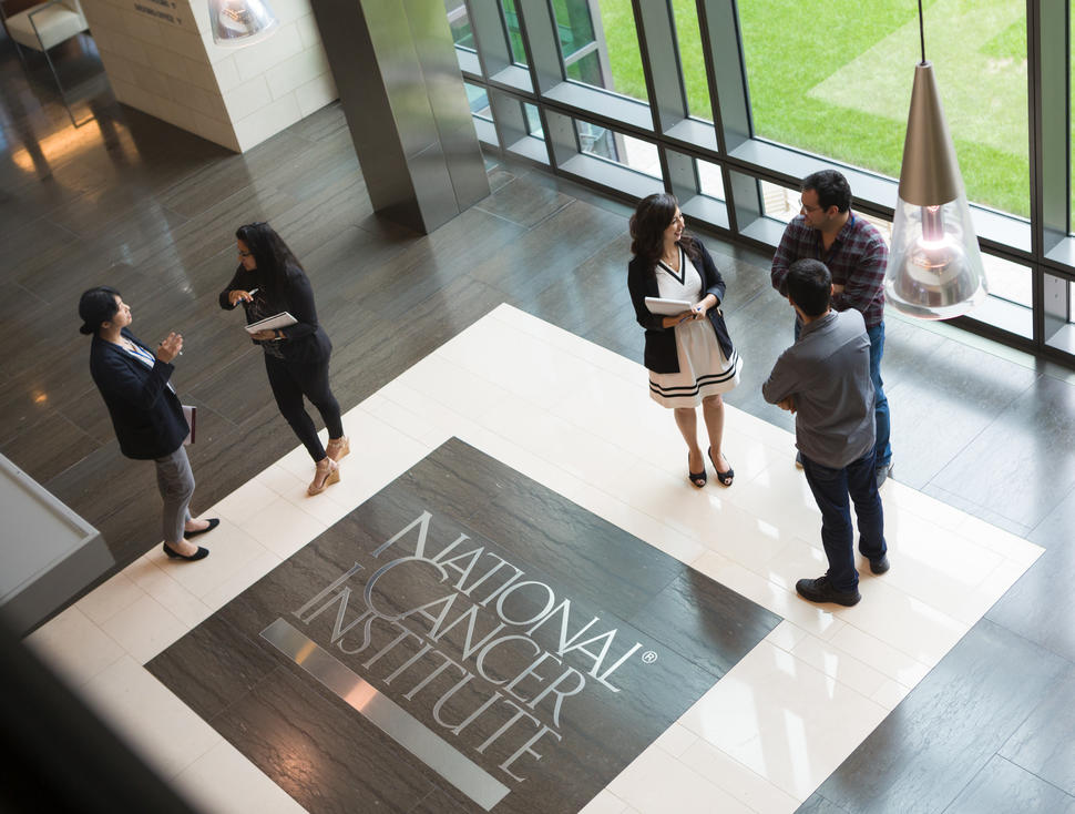 Two small groups of fellows stand talking to each other in the atrium of the NCI Shady Grove building. The NCI logo is on the floor. 