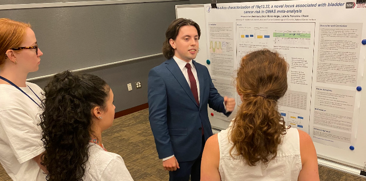 2023 summer intern in a suit stand at poster to explain his research to three listeners gathered around him.