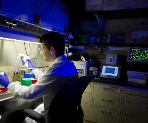 A researcher in a lab coat works at a laboratory bench, using a pipette under blue lighting, with equipment and monitors nearby.