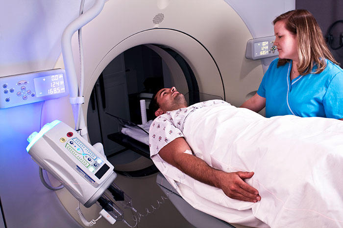 A patient lying on a bed getting ready for a CT scan with a healthcare worker in blue scrubs standing beside them for assistance
