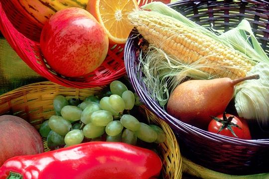 Baskets of fruits and vegetables, including an apple, an orange, an ear of corn, a pear, a tomato, a red bell pepper, and grapes.