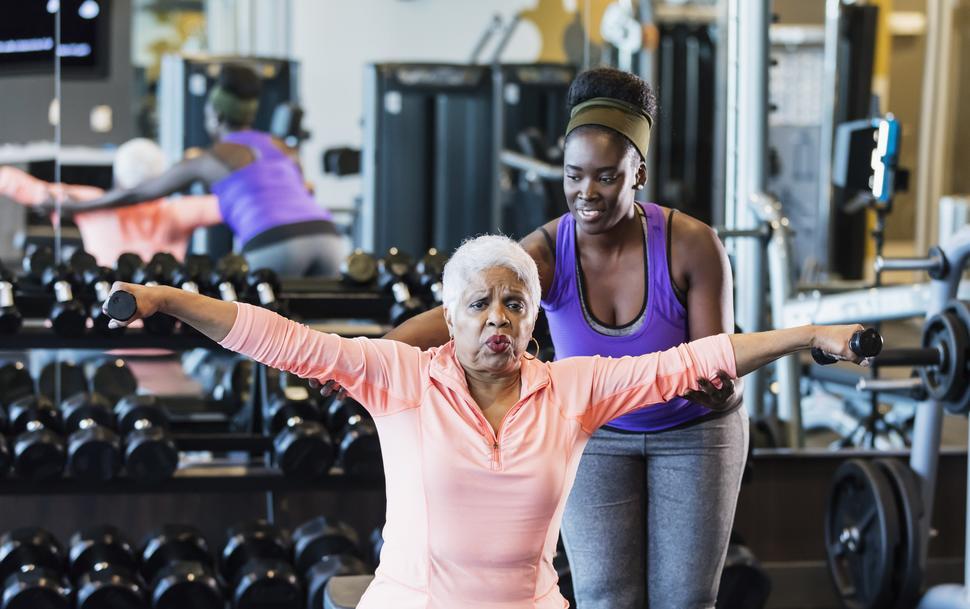 Woman in her 60s, lifting barbells up with the assistance of a trainer.