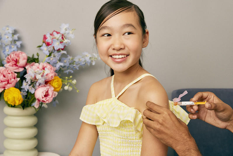 An Asian girl smiles while she receives a vaccine in her shoulder.