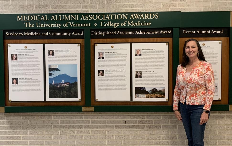 Sharon Savage stands in front of a wall of Medical Alumni Association Awards from the University of Vermont College of Medicine, including the award she received, Distinguished Academic Achievement. 