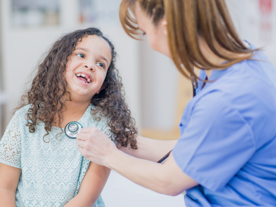 A health care provider with a ponytail puts a stethoscope on a smiling child's chest.