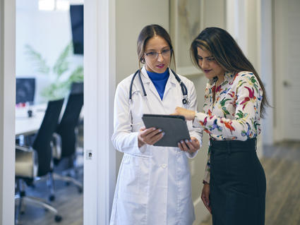 Two adult women, one a doctor and one a patient, stand in a hallway looking at a tablet. The doctor wears a lab coat over a blue top, a stethoscope around her neck, and glasses. The patient is dressed in a floral top and black skirt. The patient has a happy expression; the doctor is neutral. To the left of the doctor, an open door shows a conference room with a table, chairs, and TV. 