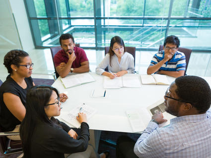 A group of people, three men and three women, dressed in business casual clothing sit around a conference table. One person, a Black man wearing glasses and a button up shirt who is closer to the camera, points at a paper on the table as the others look at him, listening intently. Other people have open notebooks or stacks of paper in front of them. The background shows glass walls, though which a flight of stairs can be shown, as well as trees outside the building, indicating a modern office.