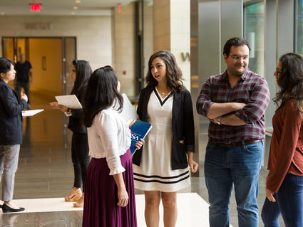 A group of six fellows - five women and one man - have conversations in a hallway. Four of the fellow are talking in the foreground; two are a further back behind them. The four fellows are having two separate one-on-one conversations. All of them are dressed in business casual clothing and have happy or neutral expressions. Floor to ceiling windows to the right of them are indicative of a modern office building. 