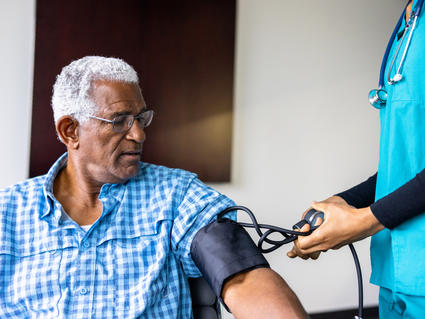 An older Black man, with white hair, glasses, and a short sleeve plaid blue shirt, looks at the blood pressure cuff on his arm. On the right of the image, a nurse in scrubs and wearing a stethoscope handles the blood pressure cuff controls.