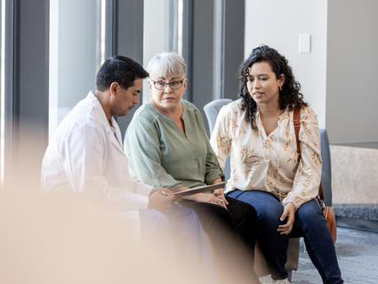 A male doctor, dressed in a white coat, looks down at a clipboard as an elderly adult female wearing glasses and her daughter, a middle-aged adult, look at the doctor. Everyone’s expression is neutral. The mother and daughter wear casual clothing. The background is indicative of a waiting area at a medical facility.