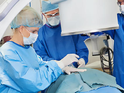 Three radiologists, one woman and two men, all dressed in blue surgical scrubs, white gloves, safety goggles and masks, stand over a patient who is covered for a procedure. A curved arm and large cylinder of the imaging machine can be seen in front of the radiologists.