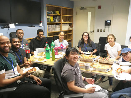 A group of young adults in casual clothing look at the camera and smile. They are seated around a conference table with multiple pizza boxes. Many of the people in the picture have a plate, some with pizza and some without. They are seated in a room with multiple TVs on the wall and bookshelves with various journals and notebooks.