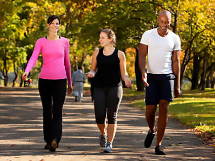 Three adults - two women and one man - dressed in casual clothing walk in a row outside. The woman in the middle is in mid-conversation with the woman next to her. Everyone is smiling. They walk on a paved pathway; in the background, green trees with yellow leaves can be seen. The shadows on the ground indicate a tree-filled park on a sunny day.