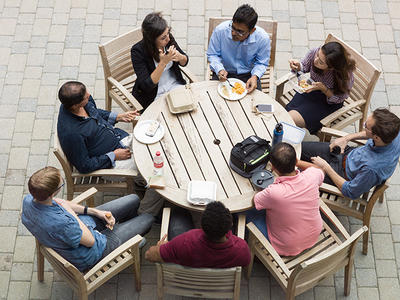 An overhead photograph of eight people, six men and two women, seated and eating lunch around a round wooden table outdoors. Everyone is dressed in business casual clothing. Some are actively eating and others are speaking or listening. The ground is made of light colored bricks. 