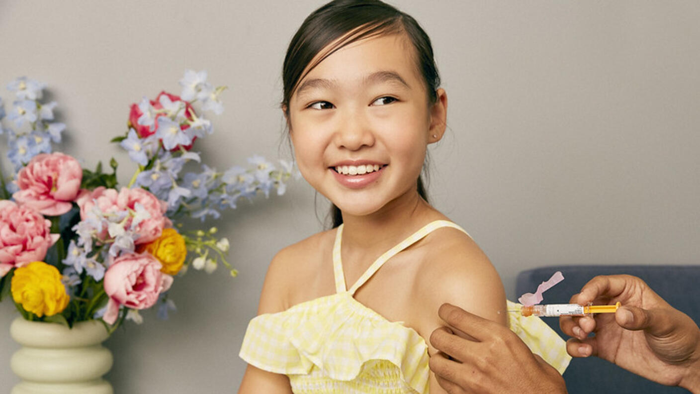 An Asian girl smiles while she receives a vaccine in her shoulder.
