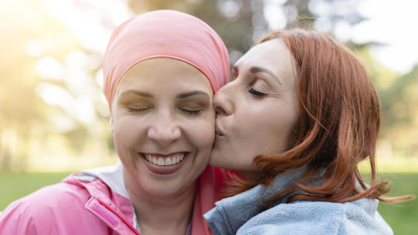 Photo of a smiling woman wearing a pink scarf on her head. Her eyes are closed and she is being embraced and kissed on the cheek by a woman with red hair.