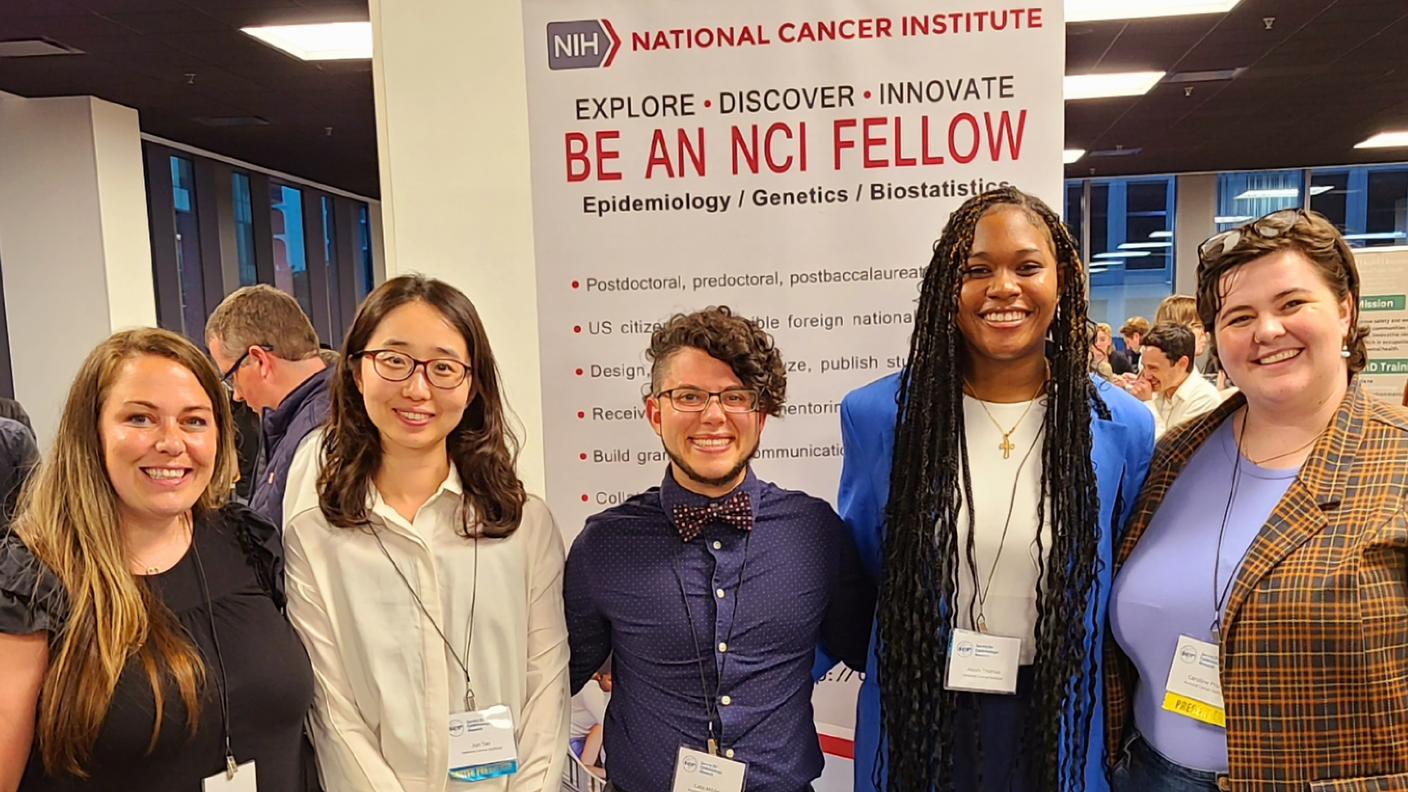 From left to right, Diane Wigfield, Jun Tao, Cato Milder, Aleah Thomas, and Caroline Pruitt stand in front of a sign that says, "Be an NCI Fellow," at the 2024 SER Annual Meeting.