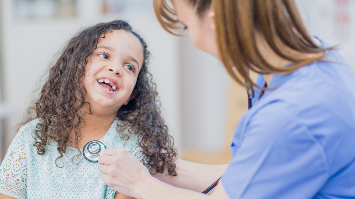 A health care provider with a ponytail puts a stethoscope on a smiling child's chest.