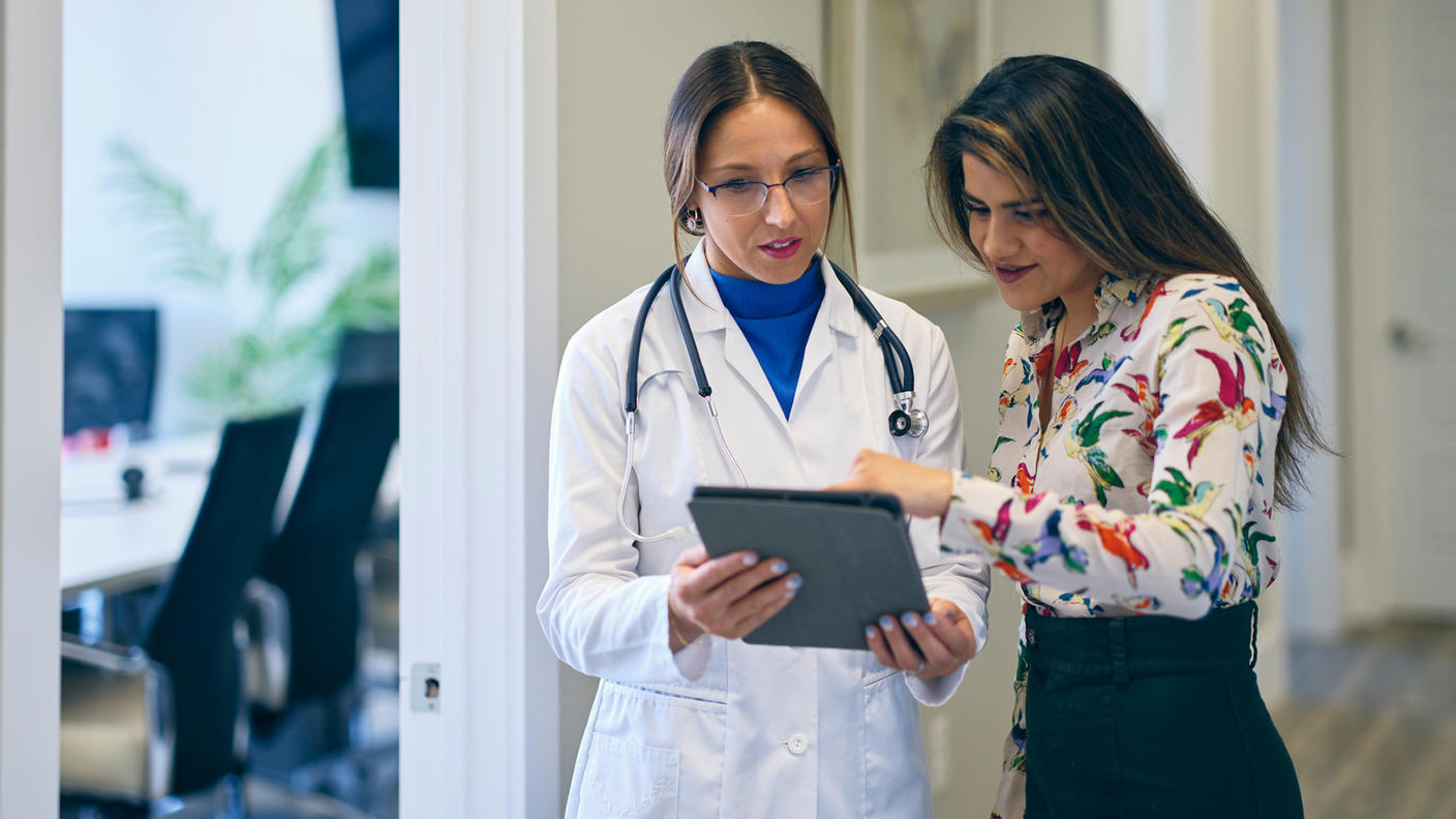 Two adult women, one a doctor and one a patient, stand in a hallway looking at a tablet. The doctor wears a lab coat over a blue top, a stethoscope around her neck, and glasses. The patient is dressed in a floral top and black skirt. The patient has a happy expression; the doctor is neutral. To the left of the doctor, an open door shows a conference room with a table, chairs, and TV. 
