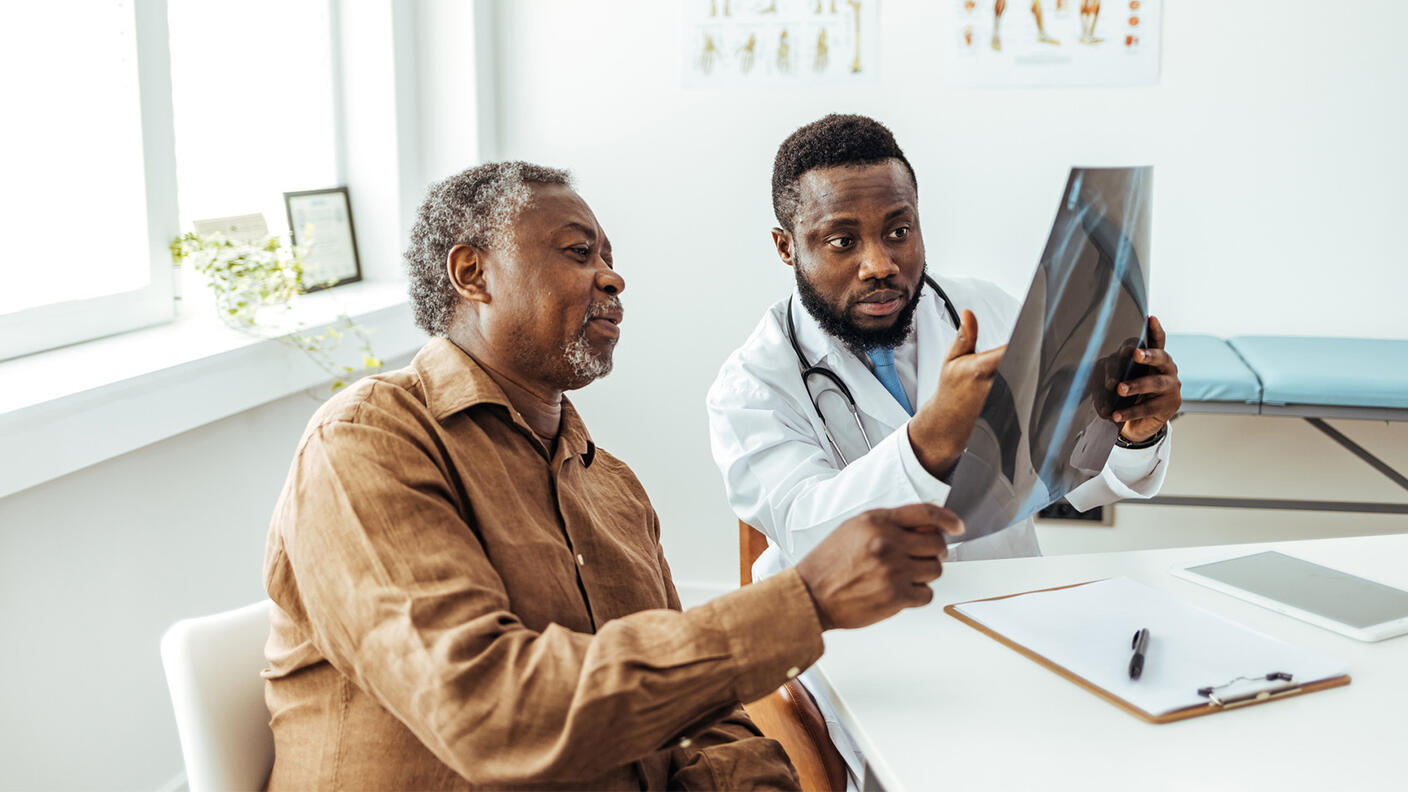 A middle-aged doctor and old patient, both Black men, examine an x-ray in the doctor’s office. The doctor wears a lab coat and stethoscope over a shirt and tie; the older man wears a casual button up brown shirt.
