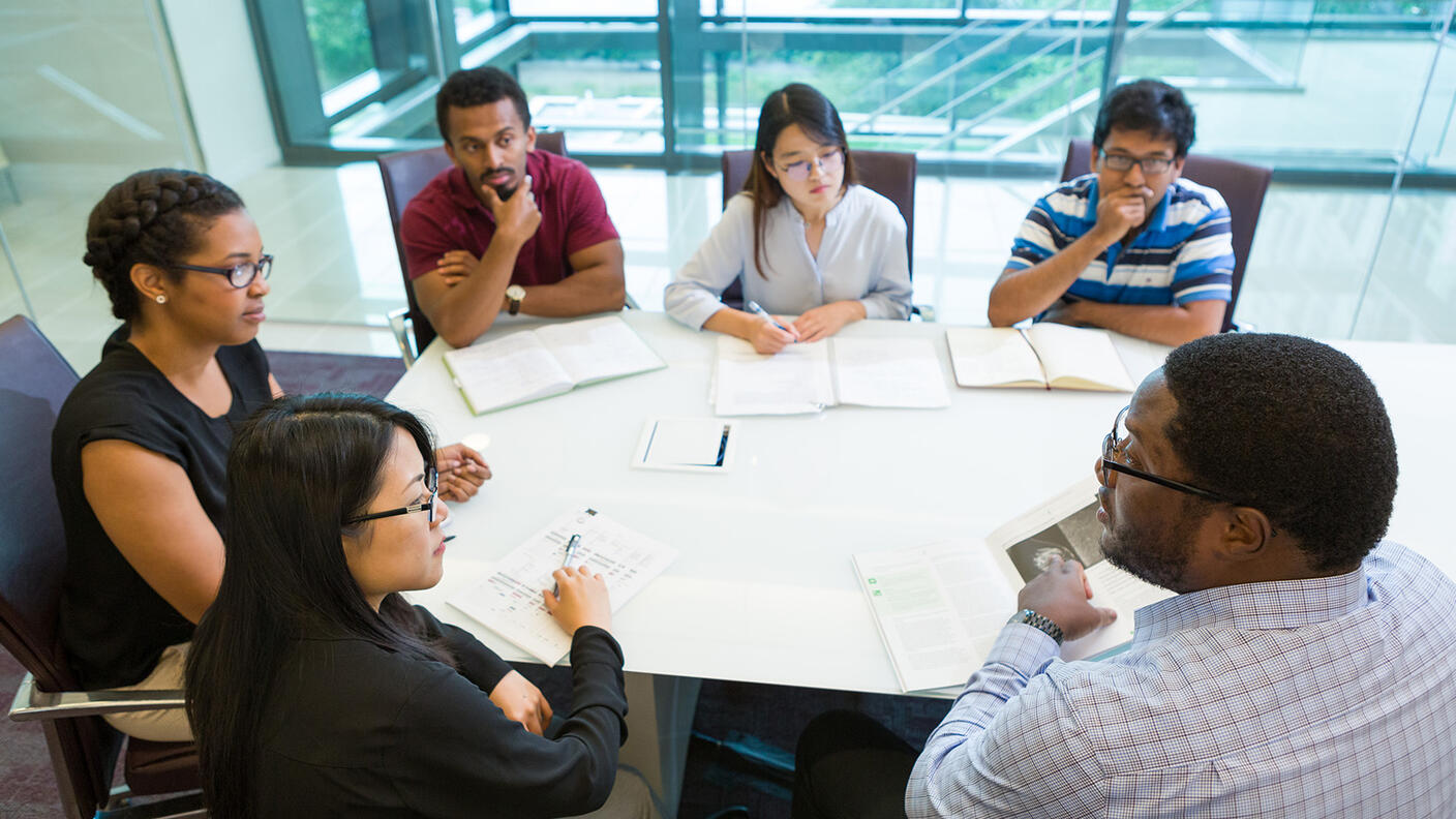 A group of people, three men and three women, dressed in business casual clothing sit around a conference table. One person, a Black man wearing glasses and a button up shirt who is closer to the camera, points at a paper on the table as the others look at him, listening intently. Other people have open notebooks or stacks of paper in front of them. The background shows glass walls, though which a flight of stairs can be shown, as well as trees outside the building, indicating a modern office.
