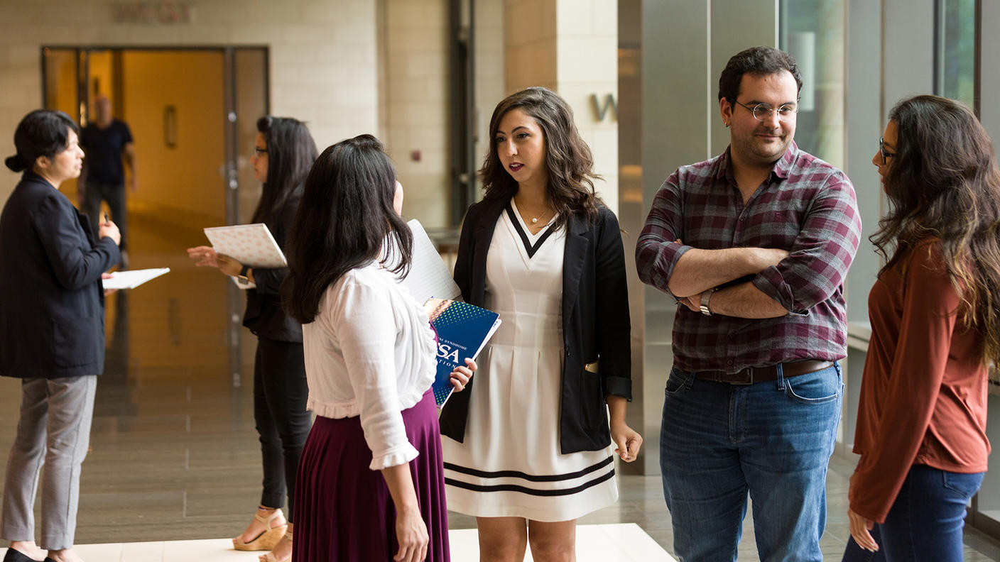 A group of six fellows - five women and one man - have conversations in a hallway. Four of the fellow are talking in the foreground; two are a further back behind them. The four fellows are having two separate one-on-one conversations. All of them are dressed in business casual clothing and have happy or neutral expressions. Floor to ceiling windows to the right of them are indicative of a modern office building. 