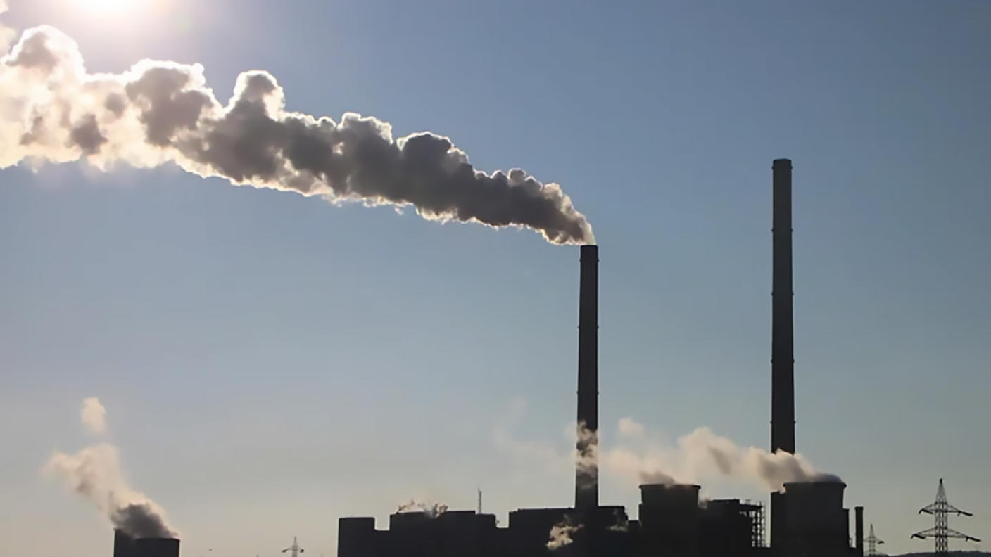 Industrial smokestacks in silhouette emit a single large stream of smoke from a tall stack with two other minor plumes of smoke from shorter stacks. In the top left corner the sun is shining, and the sky is blue and cloudless.