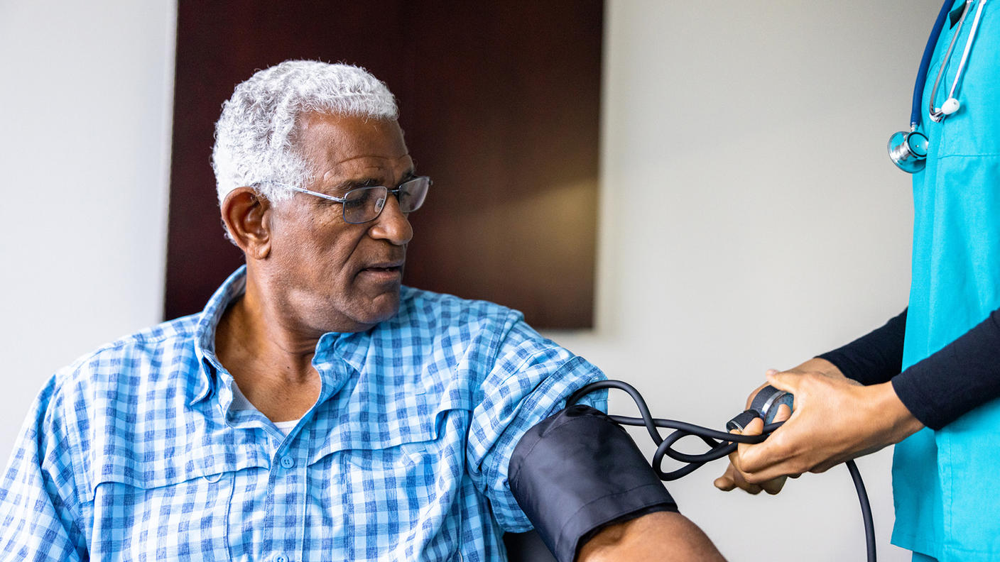 An older Black man, with white hair, glasses, and a short sleeve plaid blue shirt, looks at the blood pressure cuff on his arm. On the right of the image, a nurse in scrubs and wearing a stethoscope handles the blood pressure cuff controls.