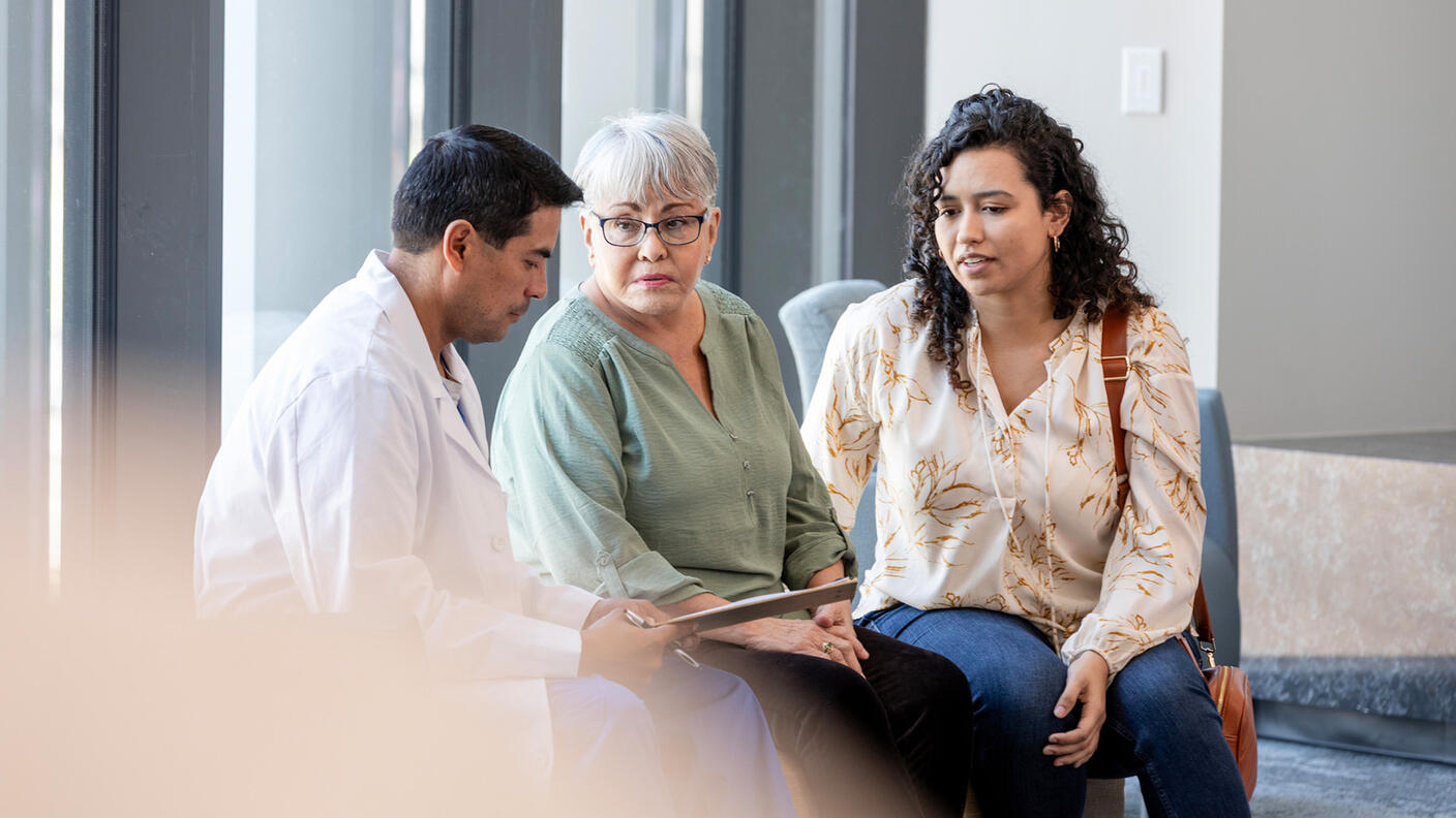A male doctor, dressed in a white coat, looks down at a clipboard as an elderly adult female wearing glasses and her daughter, a middle-aged adult, look at the doctor. Everyone’s expression is neutral. The mother and daughter wear casual clothing. The background is indicative of a waiting area at a medical facility.