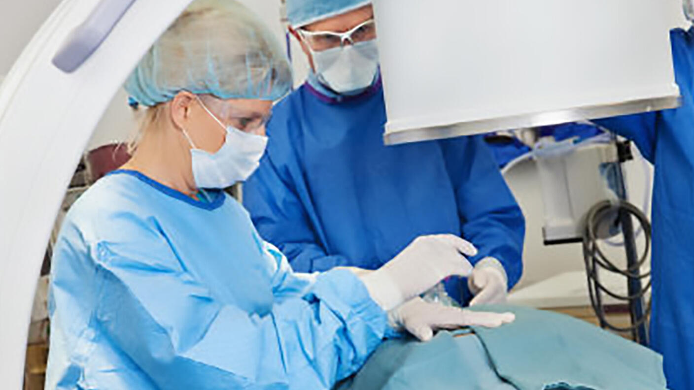Three radiologists, one woman and two men, all dressed in blue surgical scrubs, white gloves, safety goggles and masks, stand over a patient who is covered for a procedure. A curved arm and large cylinder of the imaging machine can be seen in front of the radiologists.