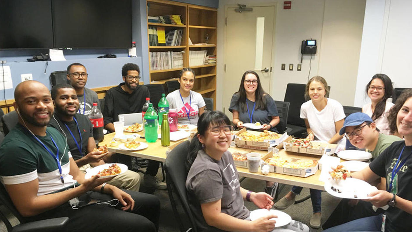 A group of young adults in casual clothing look at the camera and smile. They are seated around a conference table with multiple pizza boxes. Many of the people in the picture have a plate, some with pizza and some without. They are seated in a room with multiple TVs on the wall and bookshelves with various journals and notebooks.