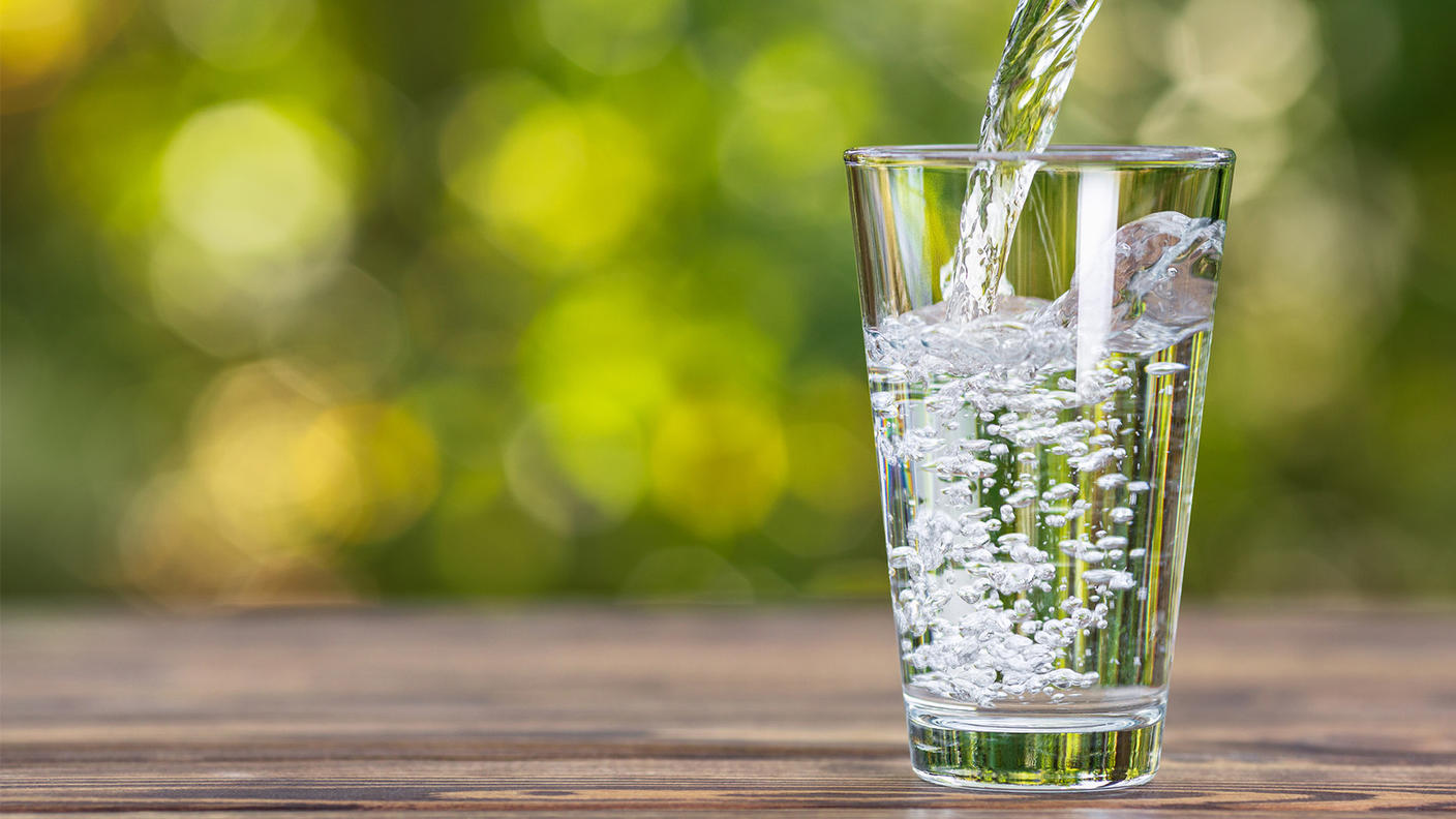 Water being poured into a clear drinking glass. The glass, which is mostly fully, is sitting on a wooden table. The pitcher can barely be seen in the right-hand corner; a healthy stream of water flows from it into the glass. In the background, a green bokeh effect can be seen, indicative of an outdoor environment on a sunny day.
