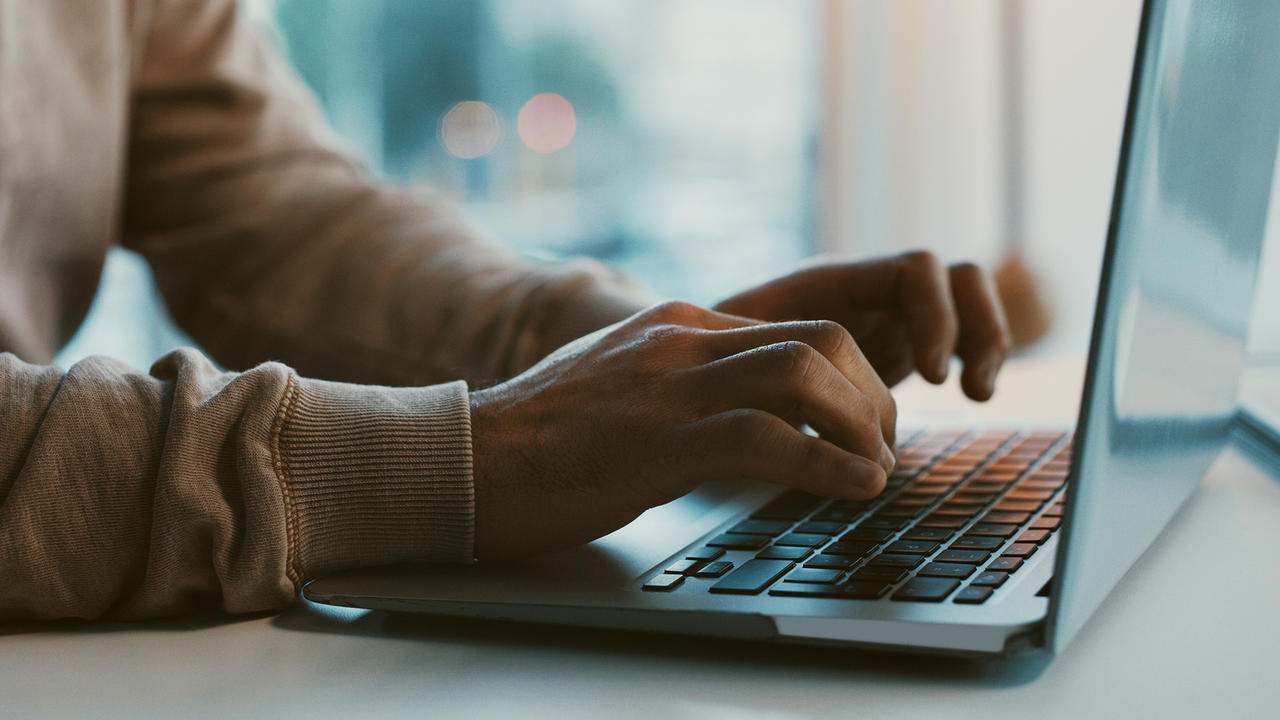 A close-up of hands typing on a laptop. The person typing is wearing a long sleeve beige sweatshirt. The laptop sits on top of a white desk. In the background, a blurry bokeh of buildings and lights can be seen, indicating the person typing is in some kind of high-rise apartment building.