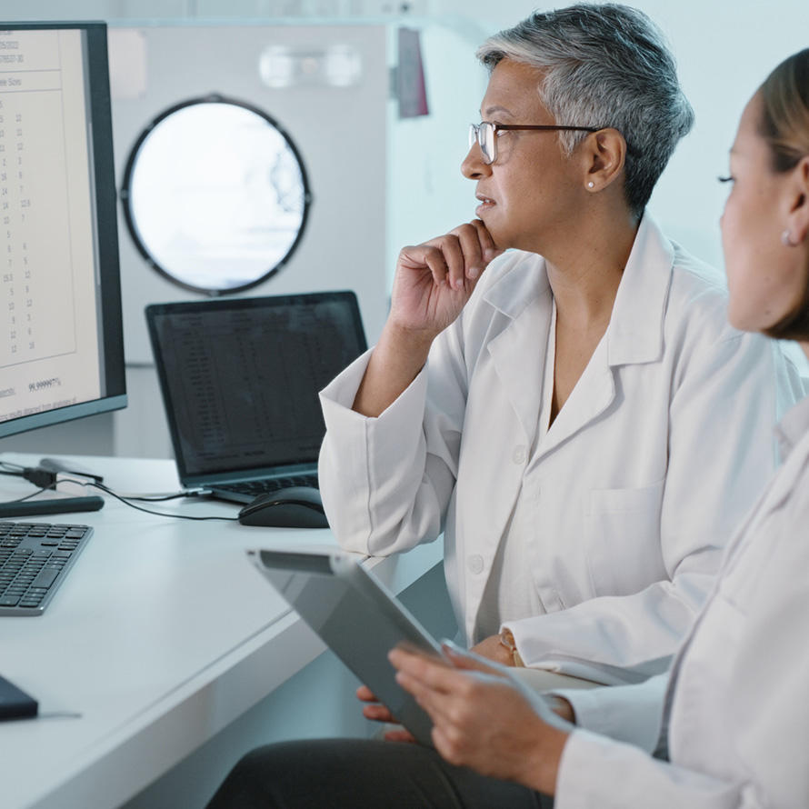 Two female scientists, one of whom is older and has grey hair and glasses, the other whom is younger with brown hair, look at a large computer monitor with data on it. The older scientist holds her hand against her chin. The younger scientist holds a tablet. The environment is mostly white, with a lamp, notebook, laptop, mouse, and keyboard on the desk. The background is indicative of a lab environment. 