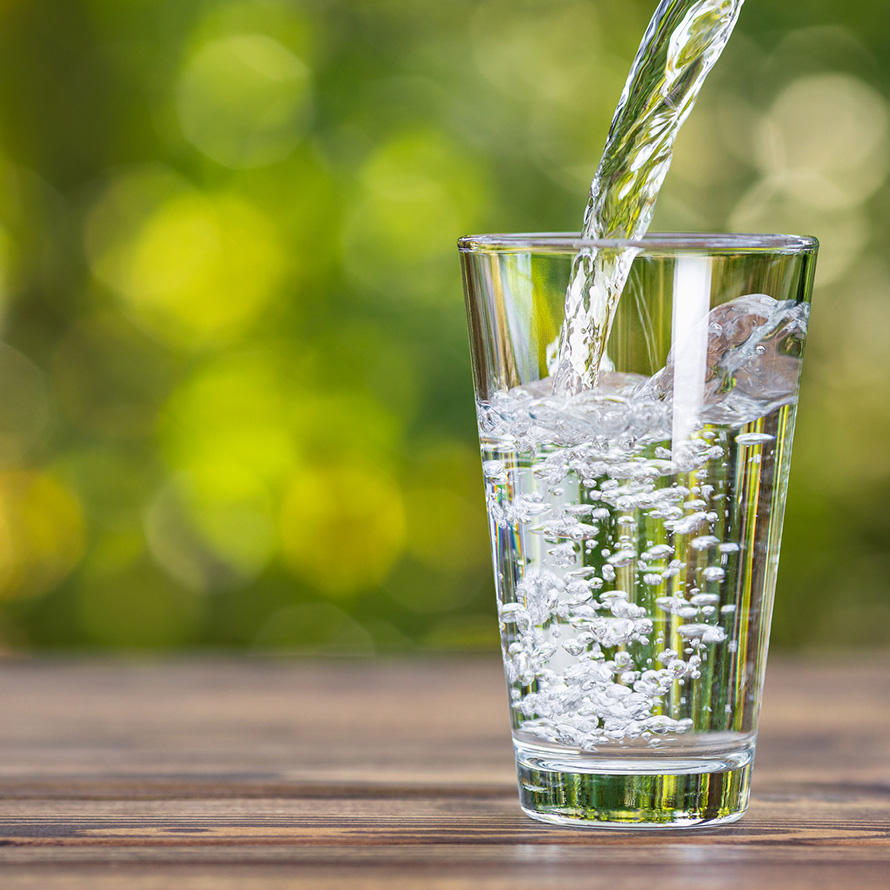 Water being poured into a clear drinking glass. The glass, which is mostly fully, is sitting on a wooden table. The pitcher can barely be seen in the right-hand corner; a healthy stream of water flows from it into the glass. In the background, a green bokeh effect can be seen, indicative of an outdoor environment on a sunny day.
