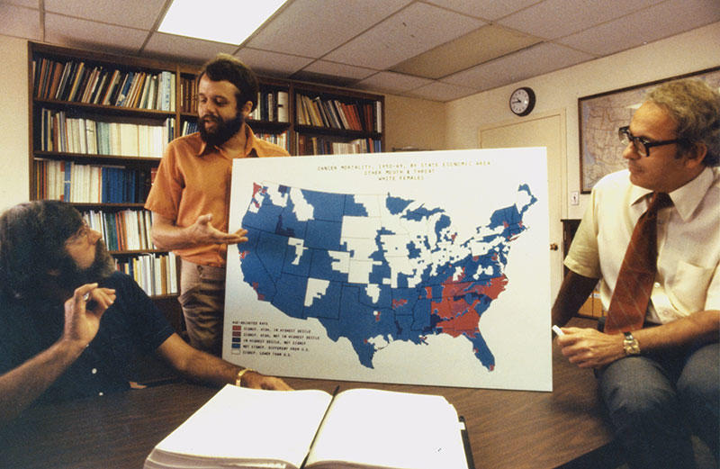 DCEG scientists look at a large board showing a map of U.S. cancer mortality in 1977