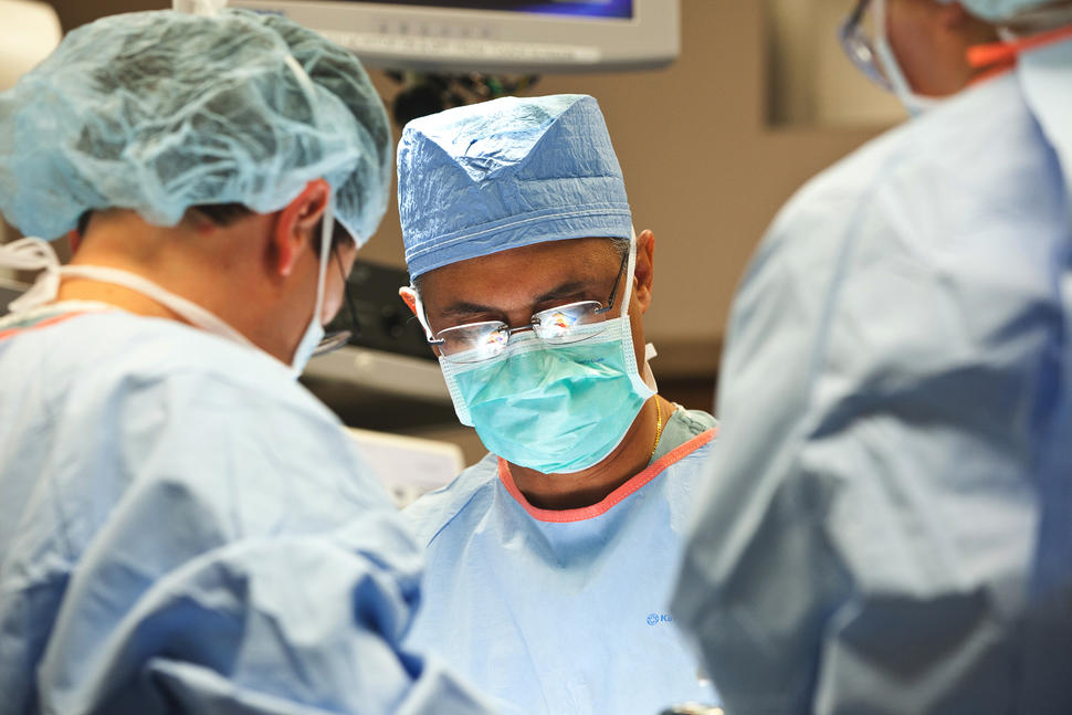 A surgeon and two assistants wearing a caps, masks, and gowns look down on a patient. There is a reflection in the surgeon's glasses.