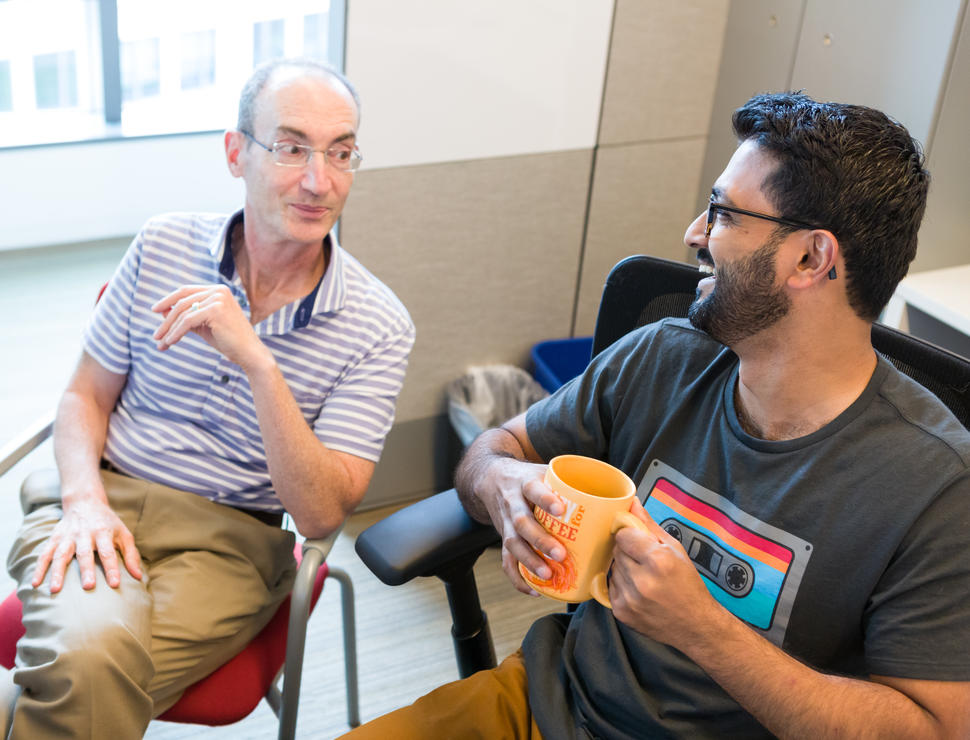 Eric Engels and his former postdoctoral fellow Parag Mahale, sit together discussing research. They are both smiling and Parag is holding a mug. 