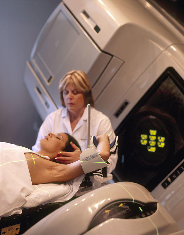 A Hispanic woman (lying on her back) is being prepared for radiation therapy by a Caucasian female radiation therapist.