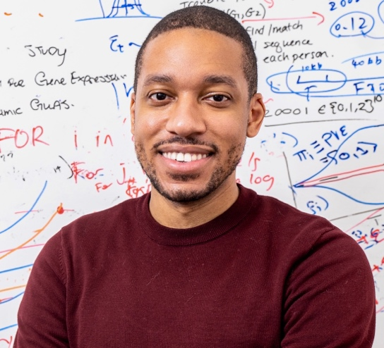 Headshot of Dr. Lorin Crawford in front of a whiteboard with notes. He is Principal Researcher at Microsoft Research New England, and Associate Professor of Biostatistics at Brown University 
