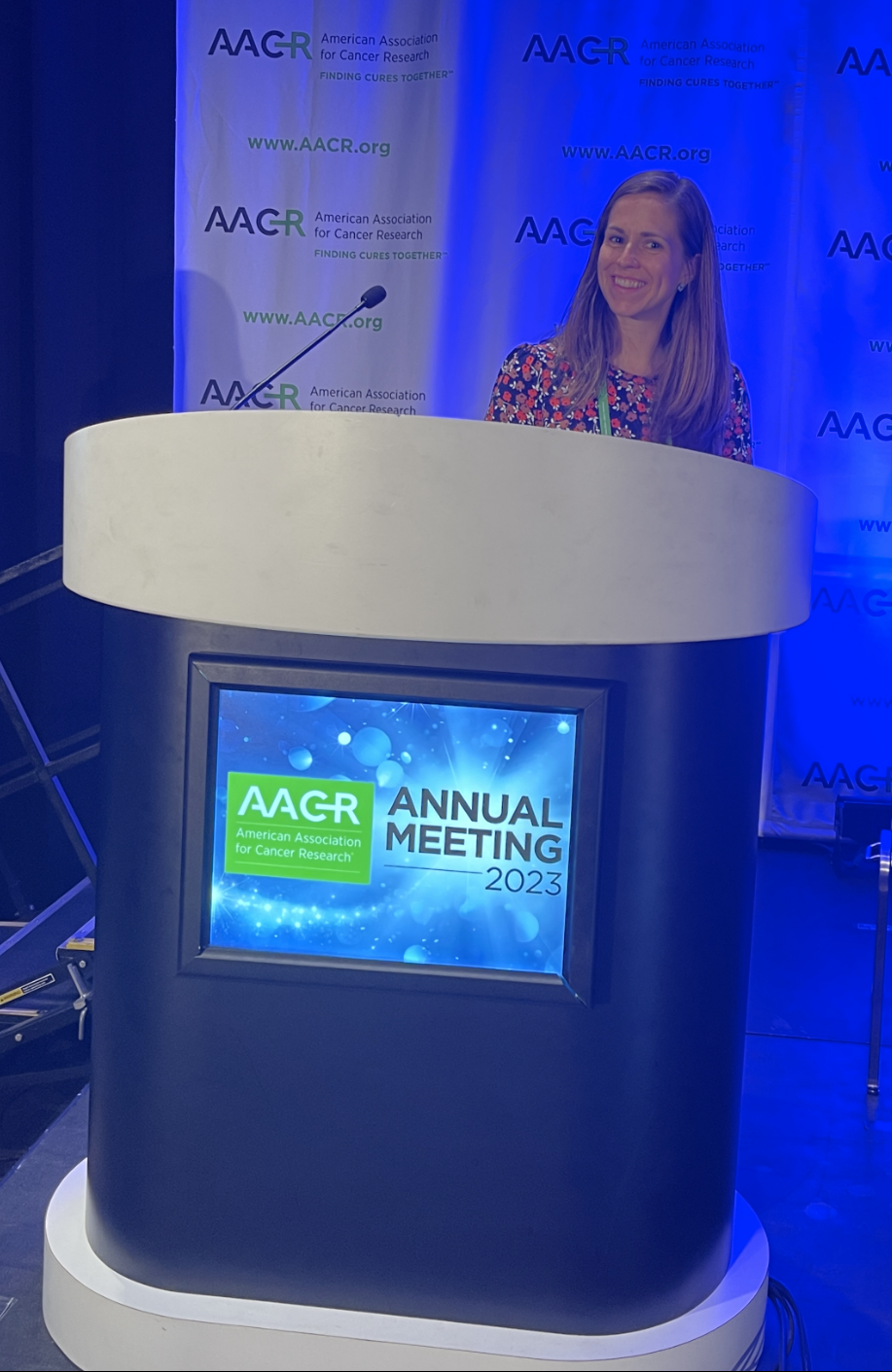 Meredith Shiels stands at a podium labeled AACR Annual Meeting 2023 at the press conference for her paper, "Opportunities for Achieving the Cancer Moonshot Goal of a 50% Reduction in Age-Adjusted Cancer Mortality by 2047."