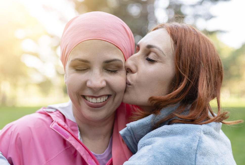 Photo of a smiling woman wearing a pink scarf on her head. Her eyes are closed and she is being embraced and kissed on the cheek by a woman with red hair.