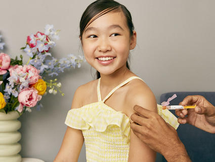 An Asian girl smiles while she receives a vaccine in her shoulder.