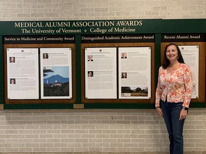 Sharon Savage stands in front of a wall of Medical Alumni Association Awards from the University of Vermont College of Medicine, including the award she received, Distinguished Academic Achievement. 