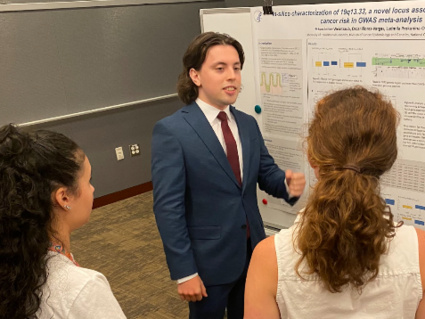 2023 summer intern in a suit stand at poster to explain his research to three listeners gathered around him.