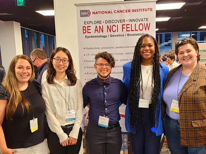 From left to right, Diane Wigfield, Jun Tao, Cato Milder, Aleah Thomas, and Caroline Pruitt stand in front of a sign that says, "Be an NCI Fellow," at the 2024 SER Annual Meeting.