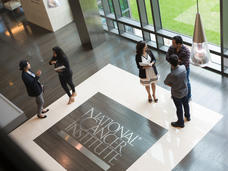 Two small groups of fellows stand talking to each other in the atrium of the NCI Shady Grove building. The NCI logo is on the floor. 